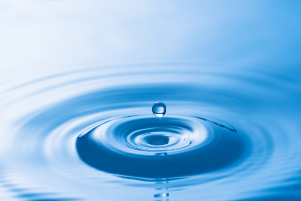 Surface of water appears blue, with a water drop creating a ripple on the surface of the water; the rings radiate out in a circle. 